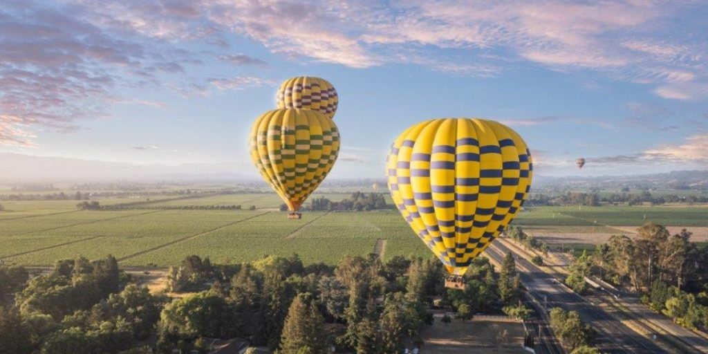 Sunrise hot air balloon ride - two yellow and blue checkered hot air balloons floating above expansive agricultural farmland and vineyards with scattered trees during early morning light with a third balloon visible in the distant sky