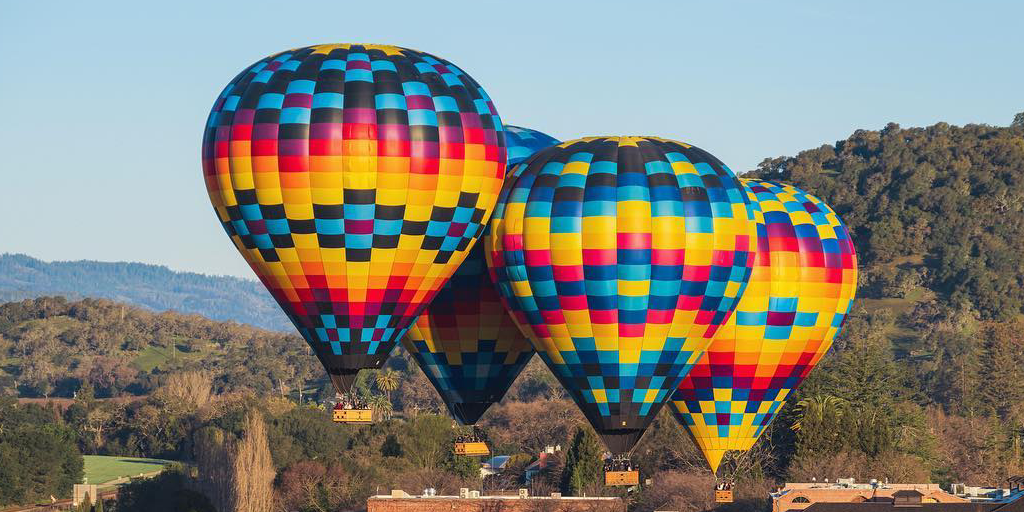 Sunrise hot air balloon ride - a blue and white striped hot air balloon drifting low over rolling vineyard-covered hills, agricultural fields, and wooded areas with a river winding through the scenic landscape during morning flight