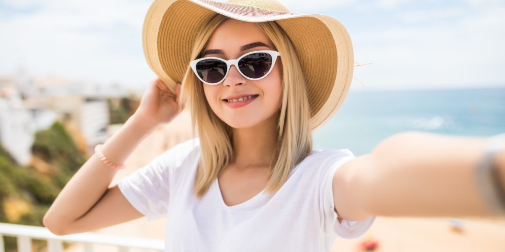 What to Wear on a Hot Air Balloon Ride in Napa Valley - a woman wearing a white t-shirt, straw sun hat, and white cat-eye sunglasses against a coastal background, displaying appropriate attire and accessories for outdoor balloon flight