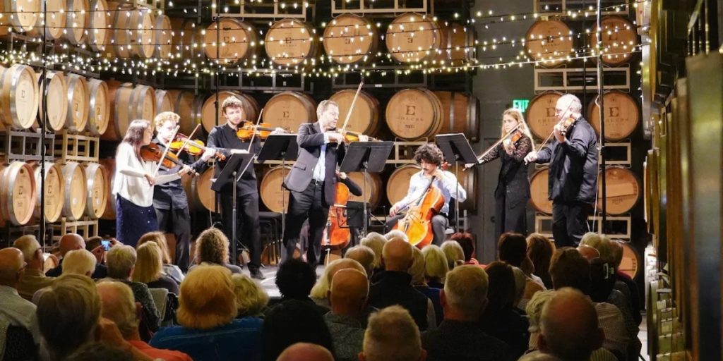Napa in August - Music in the Vineyards: a classical ensemble performs in a winery barrel room with wooden wine barrels stacked behind them and twinkling overhead lights.