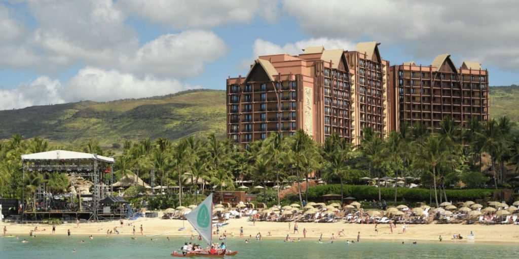 Napa in August - The Perseid Experience: a beachfront resort with Tudor-style architecture nestled against green hills, with a sandy beach, ocean, and event stage below.