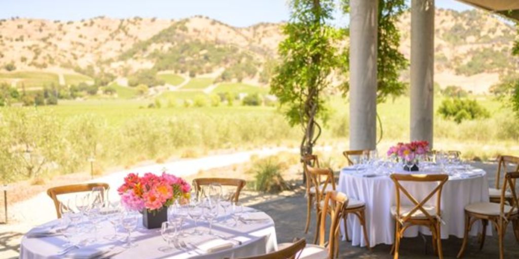Napa in August - Poetry Winemaker Dinner: an elegant outdoor dining setup with white-draped tables, wooden chairs, vibrant floral centerpieces, and sweeping vineyard views.