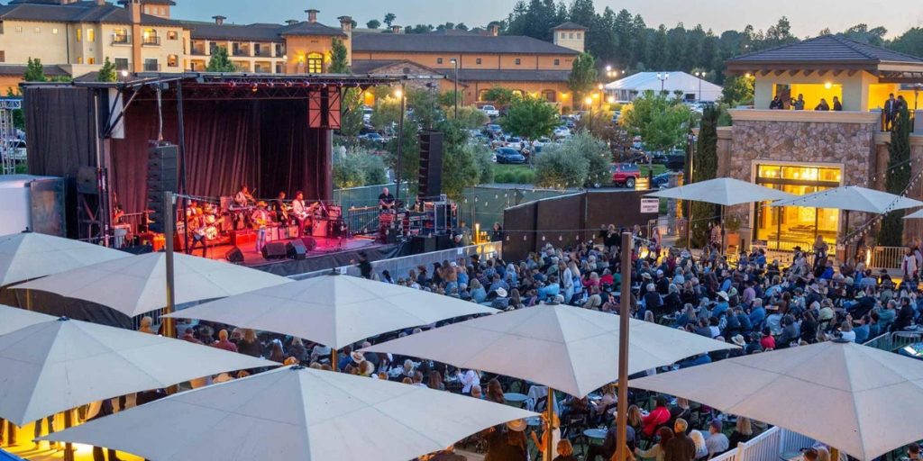 Blue Note Napa Summer Sessions outdoor concert featuring a live band performing on an illuminated stage with a large crowd of spectators seated under white tent canopies in a vineyard venue setting.