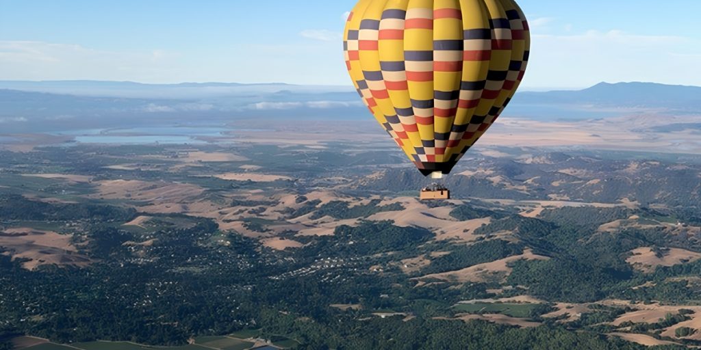 Napa Valley Aloft sunrise balloon flight – colorful hot air balloon floating above scenic vineyard landscape with rolling hills, forests, and mountains during early morning aerial tour